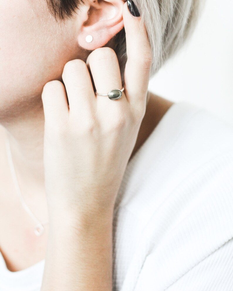 May include: A close-up of a person's hand wearing a silver ring with a dark green oval-shaped stone. The person is also wearing a small silver stud earring in their left ear.