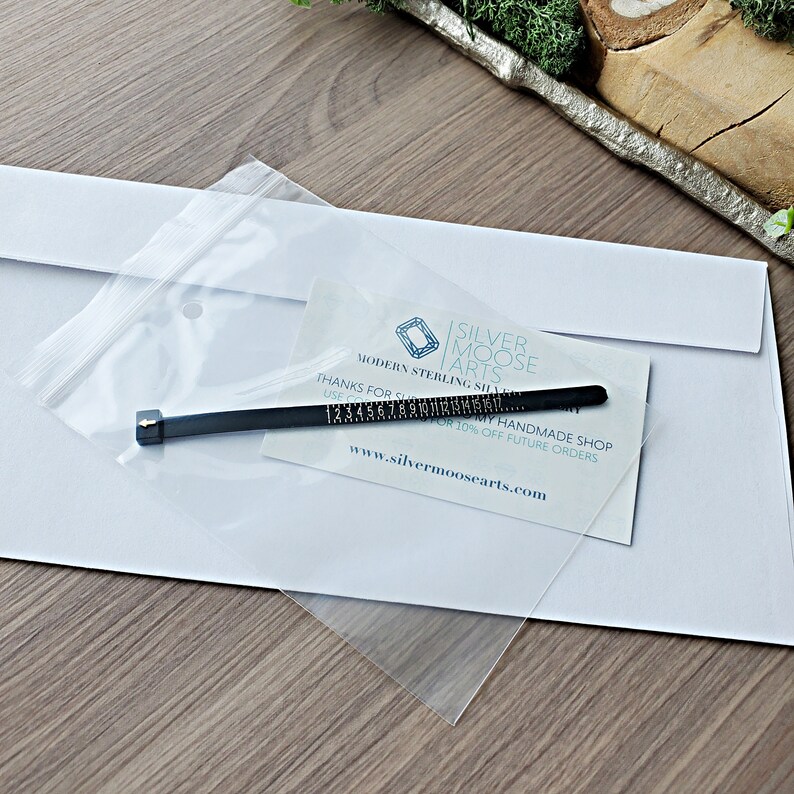 May include: A white envelope with a clear plastic bag containing a black ring sizer with markings from 1 to 16. The bag also contains a business card with the text "Silver Moose Arts" and "Modern Sterling Silver Jewelry Handmade Shop" and the website address "www.silvermoosearts.com".