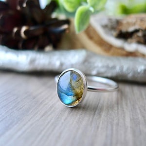 May include: A silver ring featuring a blue and green labradorite gemstone. The ring has a simple band and the stone is oval shaped.