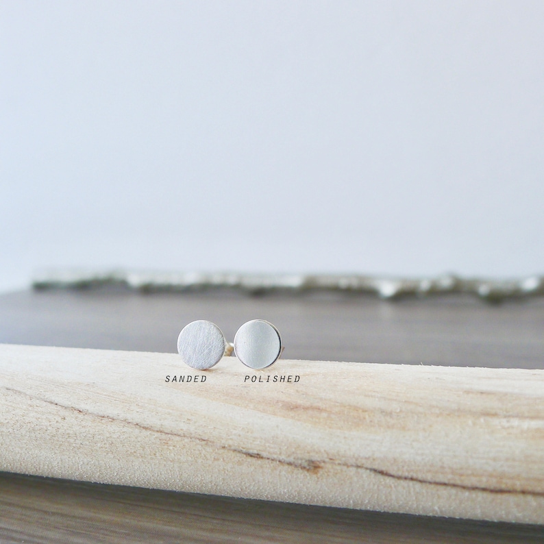 May include: Two silver round earrings, one with a sanded finish and the other with a polished finish. The earrings are displayed on a wooden surface.
