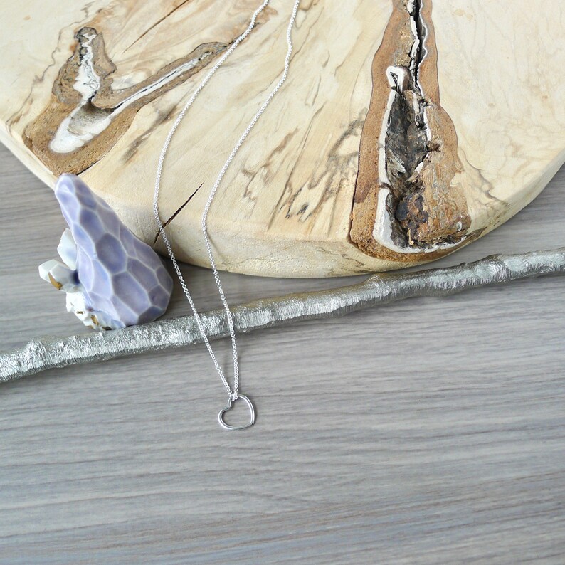 May include: A silver chain necklace with a simple open heart pendant. The necklace is laying on a wooden surface.