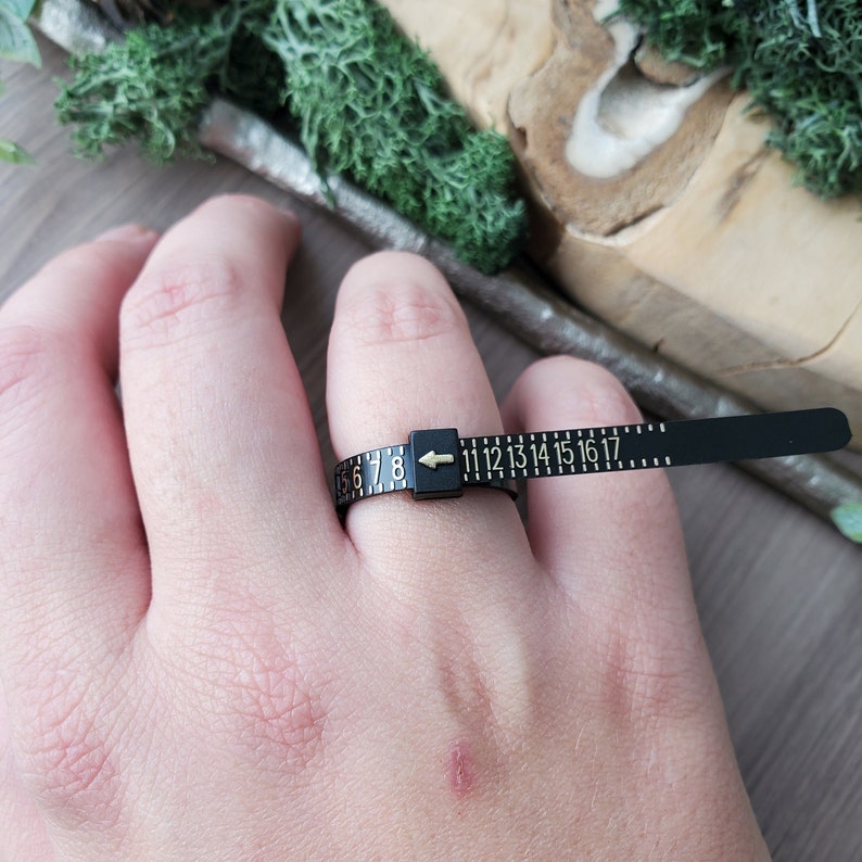 May include: A black ring sizer with gold numbers and an arrow. The sizer is on a finger and shows sizes 5 through 17.
