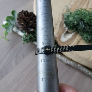 May include: A black ring sizer with gold numbers and an arrow pointing to the number 9. The sizer is wrapped around a metal ring mandrel.