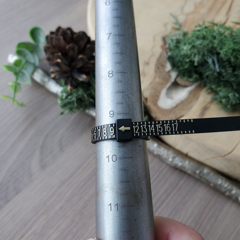 May include: A black ring sizer with gold numbers and an arrow pointing to the number 9. The sizer is wrapped around a metal ring mandrel.