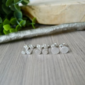 May include: A set of four silver stud earrings with a brushed finish. The earrings are arranged in a row on a wooden surface.