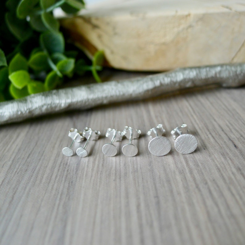 May include: A set of four silver stud earrings with a brushed finish. The earrings are arranged in a row on a wooden surface.