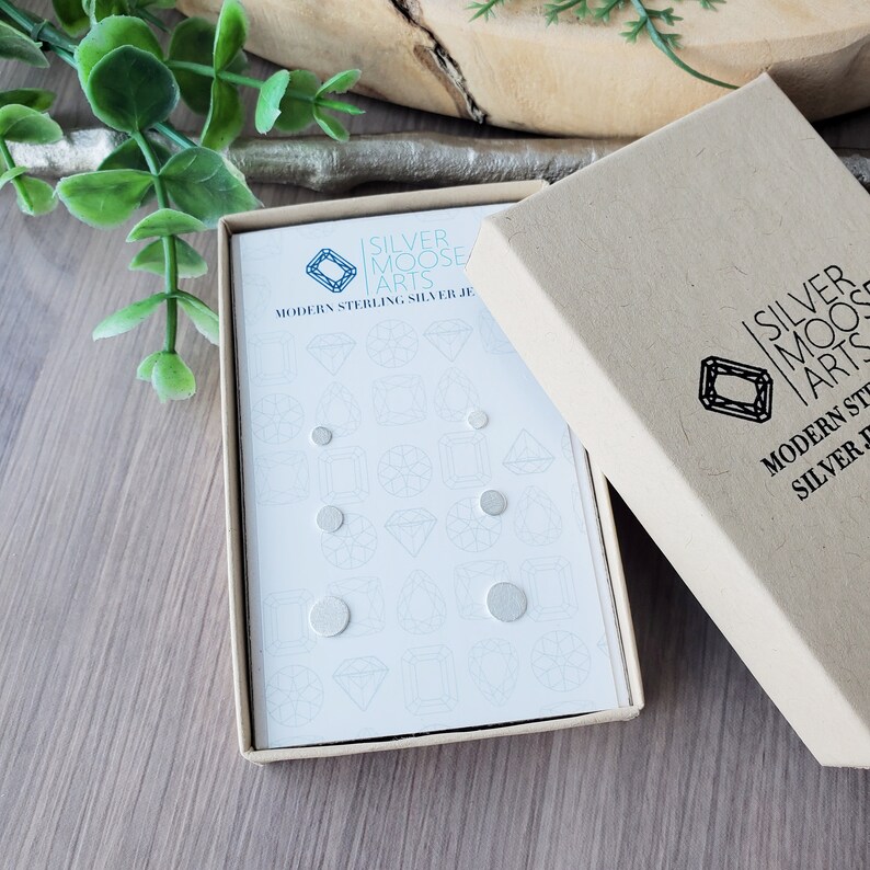 May include: A white box with a blue logo that says "Silver Moose Arts" and the text "Modern Sterling Silver Jewelry". The box contains a set of silver stud earrings.
