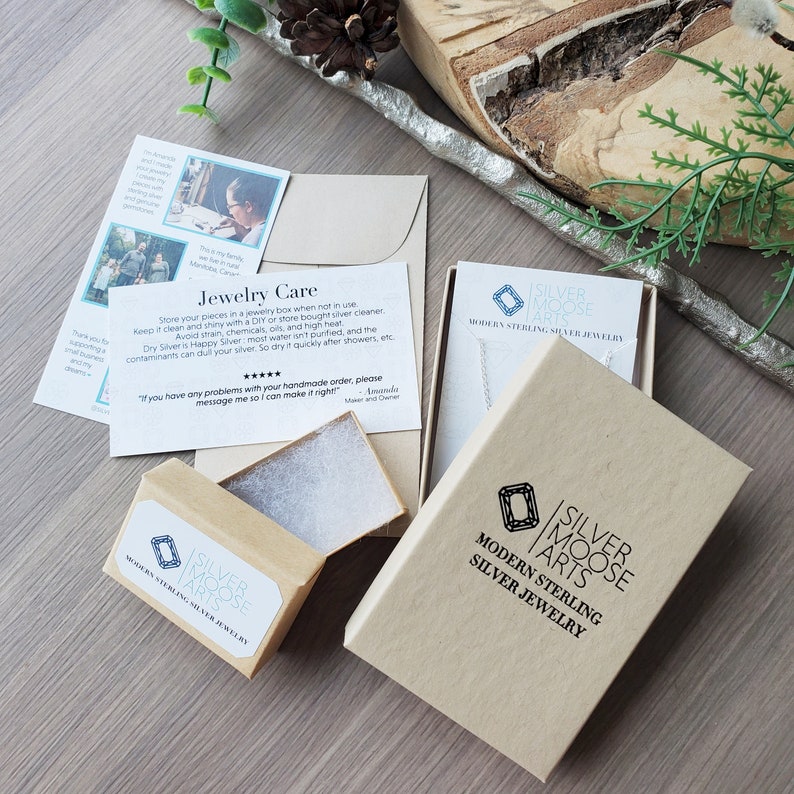 May include: A brown cardboard box with a white label that says "Silver Moose Arts Modern Sterling Silver Jewelry". The box is open and there is a white foam insert inside. The box is sitting on a wooden table.