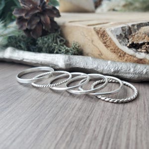 May include: A set of silver rings with different designs, including plain and twisted bands. The rings are displayed on a wooden surface, with a pine cone and moss visible in the background. The rings are made of silver.