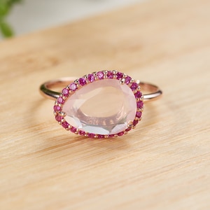 May include: Rose gold ring featuring a pear-shaped, light pink gemstone surrounded by small, round, magenta-colored stones. The ring is set on a light-colored wooden surface, showcasing the jewelry's design and color contrast.
