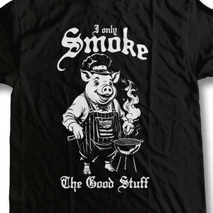 May include: Black t-shirt with a white graphic of a pig wearing a chef's hat and apron, grilling on a barbecue. The text "I only Smoke The Good Stuff" is printed below the pig.