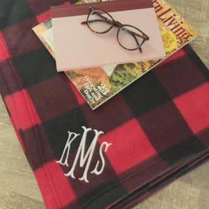 May include: A red and black buffalo check fleece blanket with the monogram "KMS" embroidered in white. The blanket is folded and lying on a wooden surface with a magazine and a pair of glasses on top of it.