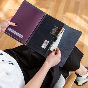 Könnte beinhalten: Offenes marineblaues Leder-Portfolio mit einem weinroten Notizbuch. Das Portfolio hat einen Stift, ein kleines goldfarbenes Dekorationselement und eine kleine Karte. Das Portfolio ist geöffnet und zeigt die Innentaschen und den Inhalt.