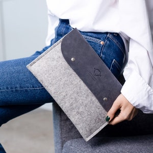May include: A grey felt and dark grey leather laptop sleeve with a logo that says "Joy & Grace" in white.