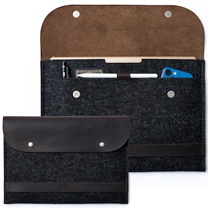 May include: A gray felt laptop sleeve with a brown leather flap and a brown leather strap. The sleeve has a small pocket on the front for a phone or other small items. The sleeve is open and shows a white tablet and a blue phone inside.