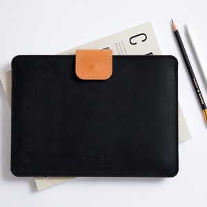 May include: Black leather laptop sleeve with a brown leather flap closure. The sleeve has a subtle embossed logo.