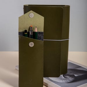 May include: An olive green leather pen case with a magnetic clasp, containing a pencil and pens. A matching journal with a white elastic closure is behind the case. A silver pencil sharpener and a book are in the foreground.