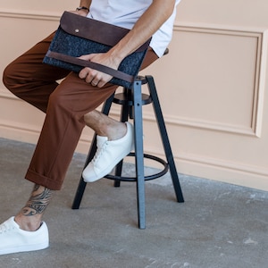 Young man sitting on the chair nice dressed, with white sneakers. Tatoo on the leg. A man is holding leather and felt laptop case.