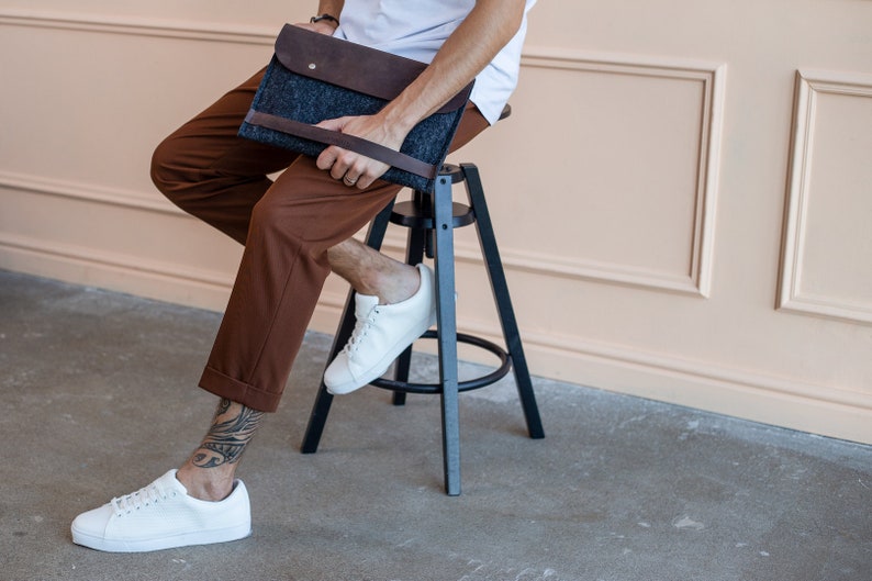 Young man sitting on the chair nice dressed, with white sneakers. Tatoo on the leg. A man is holding leather and felt laptop case.