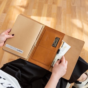 Può includere: Diario in pelle marrone chiaro aperto, con interno marrone e una carta. Il diario è aperto, rivelando un taccuino beige e una tasca in pelle marrone con un logo dorato. È visibile anche una penna.