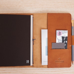 May include: A brown leather notebook cover with a black notebook inside. The cover has a pocket on the right side with a card and a pencil. The cover has the text "CITYSHEEP" and a logo on the bottom right corner.