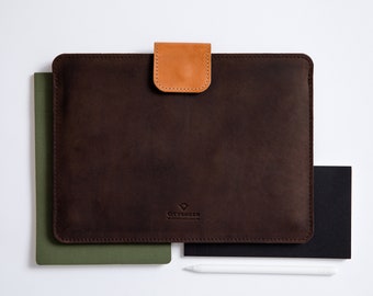 May include: A brown leather laptop sleeve with a tan leather flap closure. The sleeve has a stitched logo that reads "CITYSHEEP" in white text. The sleeve is sitting on top of a green notebook and a black notebook.