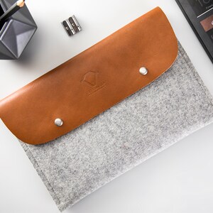 May include: A gray felt pencil case with a brown leather flap. The flap has a small logo and two silver snaps. The case is lying on a white surface.