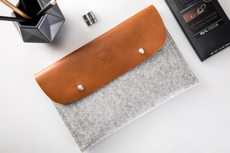 May include: A gray felt pencil case with a brown leather flap. The flap has a small logo and two silver snaps. The case is lying on a white surface.