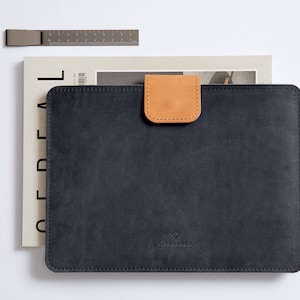 Può includere: Una custodia per laptop in pelle nera con chiusura a lembo in pelle marrone. La custodia ha un bordo cucito e il nome del marchio "CITYSCENED" è in rilievo sul davanti.