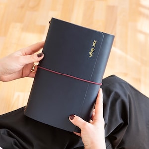 Könnte beinhalten: Ein marineblaues Notizbuch mit dem goldenen Text "Just Begin" wird von einer Person gehalten. Das Notizbuch ist mit einem weinroten Gummiband gesichert. Die Person trägt eine schwarze Hose und hat dunklen Nagellack.