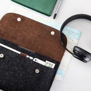 May include: A gray felt pouch with a brown felt lining, open to reveal a white stylus, a small electronic device, and a dollar bill. A pair of black over-ear headphones and a map of New York City are also visible.