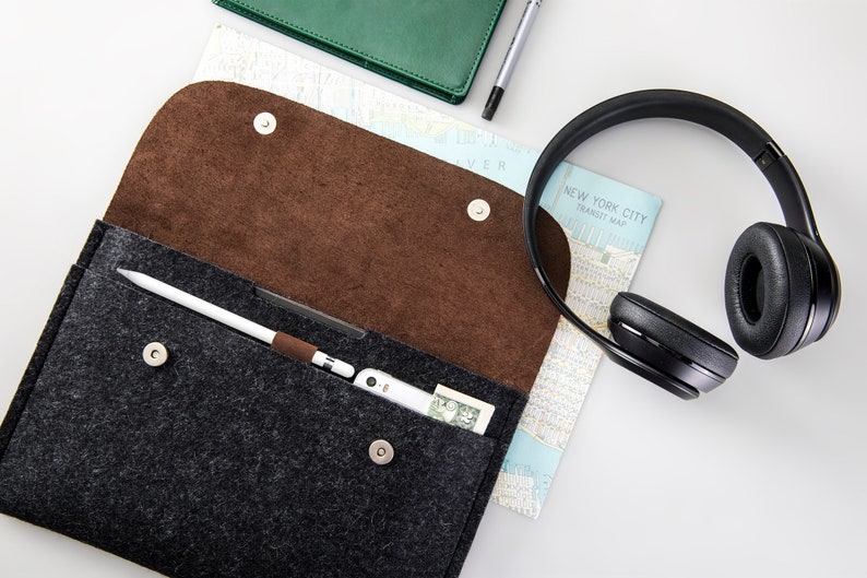 May include: A gray felt pouch with a brown felt lining, open to reveal a white stylus, a small electronic device, and a dollar bill. A pair of black over-ear headphones and a map of New York City are also visible.