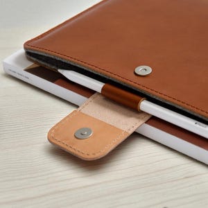 May include: A brown leather tablet case with a magnetic closure, holding a white stylus. The case is open, revealing a gray interior. A small, light brown leather holder secures the stylus. The case rests on a white book.