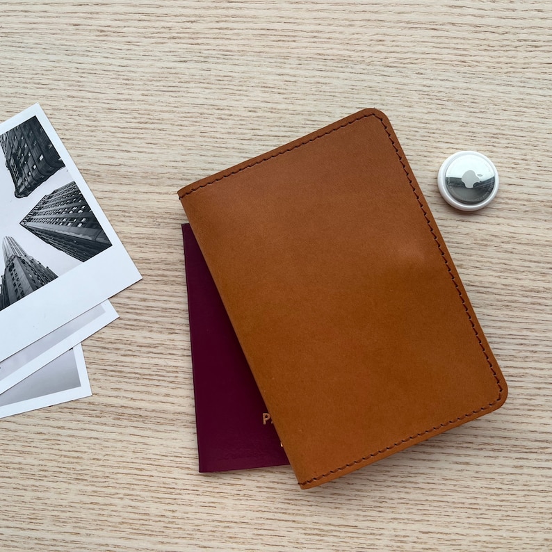 May include: A brown leather passport holder with a stitched edge. The holder is lying on a wooden surface next to a burgundy passport cover.