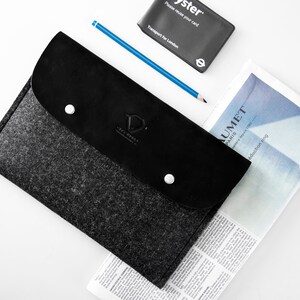 May include: A black felt and leather laptop sleeve with a silver button closure and the text "JAX HENRY" embossed on the front. The sleeve is lying on top of a newspaper.