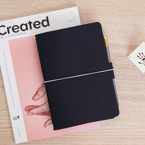 May include: A black leather-bound journal with a white elastic band closure and a gold pen. The journal is lying on top of a magazine with the text "Created in Lithuania".