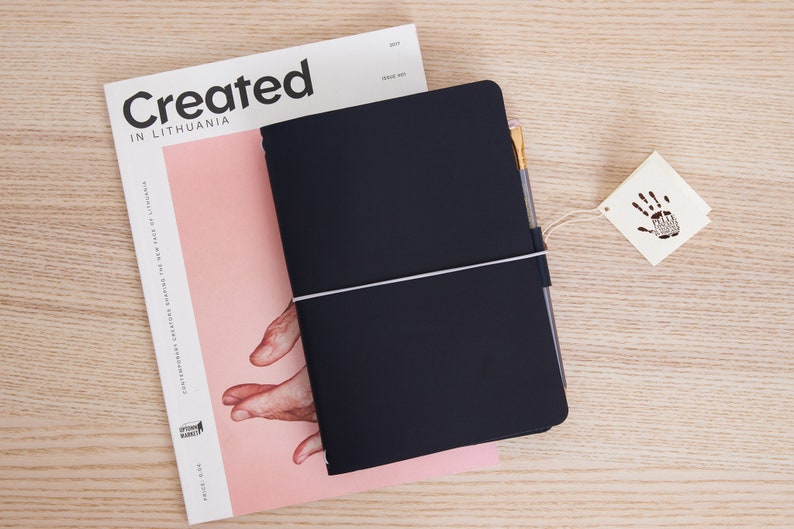 May include: A black leather-bound journal with a white elastic band closure and a gold pen. The journal is lying on top of a magazine with the text "Created in Lithuania".