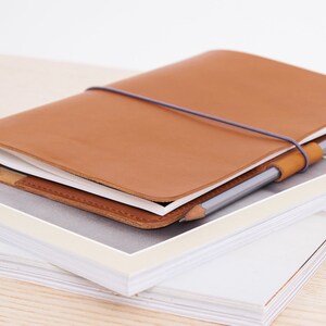 May include: A brown leather notebook with a grey elastic band and a pencil. The notebook is closed and sitting on top of a stack of white papers.