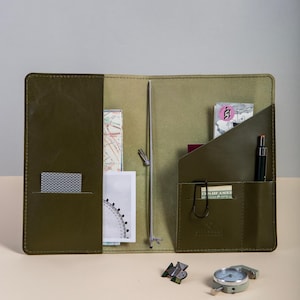 Könnte beinhalten: Olivgrünes Leder-Reisetagebuch mit mehreren Taschen, die Karten, Karten und einen Stift enthalten. Ein Kompass, ein Messgerät und eine Anstecknadel befinden sich auf der Oberfläche. Das Tagebuch ist geöffnet und zeigt einen hellgrünen Innenraum.