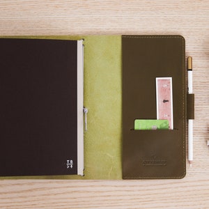May include: A brown leather notebook with a green leather cover and a pen holder. The notebook has a pocket on the right side for cards and a ruler. The notebook is open and the pages are blank.