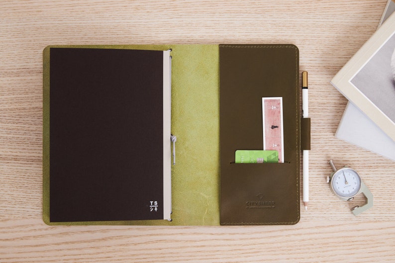 May include: A brown leather notebook with a green leather cover and a pen holder. The notebook has a pocket on the right side for cards and a ruler. The notebook is open and the pages are blank.