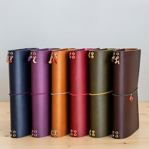 May include: Six vertical leather journals in navy, purple, tan, red, olive green, and dark brown. Each journal features gold-toned metal eyelets, a colored elastic closure band, and a small decorative charm attached to the top binding.