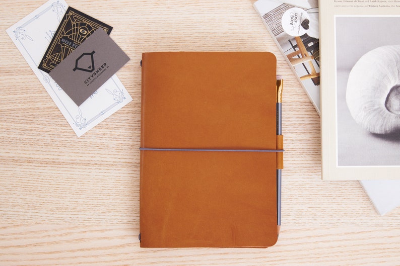May include: A brown leather bound journal with a grey elastic band and a gold pen sticking out of the top. The journal is sitting on a wooden surface.