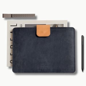 May include: A navy blue leather tablet case with a tan leather tab closure. A metal ruler and a black stylus pen are also present. The case is on top of a magazine.