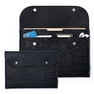 Puede incluir: Una funda para portátil de fieltro negro con una solapa de cuero negro y un bolsillo de cuero negro más pequeño en la parte delantera. La funda tiene un forro de fieltro gris y cierre de cremallera. La funda se muestra con un teléfono, un lápiz óptico y auriculares en su interior.