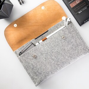 May include: A gray felt clutch with a brown leather flap. The clutch is open and has a smartphone, a stylus, and a pair of white earbuds inside.
