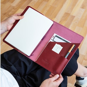 Pode incluir: Uma capa de caderno em couro vermelho escuro com design aberto, um inserto de papel branco, um cordão elástico roxo e um bolso interno contendo um pequeno cartão branco, um bloco de notas em preto e branco e uma caneta metálica.
