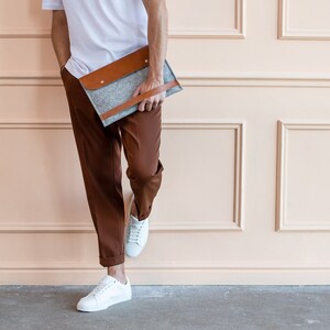 A young man is holding a leather and felt laptop case. Standing by the wall.