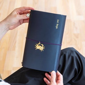 Könnte beinhalten: Ein marineblaues Notizbuch mit dem goldenen Schriftzug „Just Begin“. Ein goldener Krebs-Anhänger hängt an einem lila Gummiband. Das Notizbuch ist geöffnet und zeigt die Innenseiten.