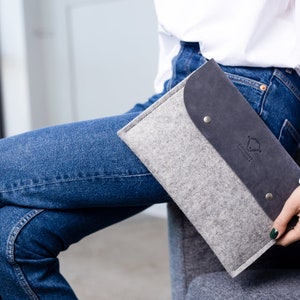 May include: A gray felt laptop sleeve with a black leather flap and a silver metal button. The sleeve has a logo that says "Simple & Sustainable".
